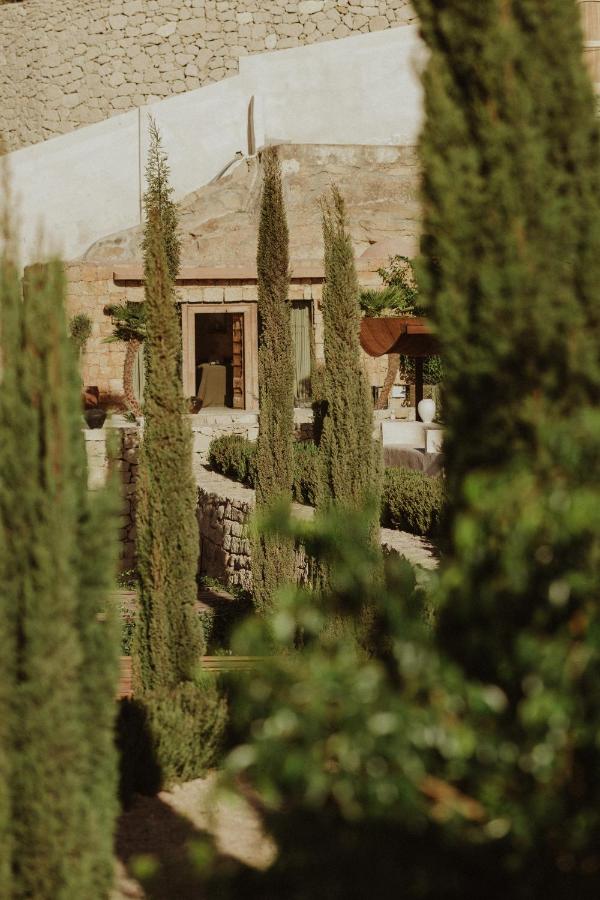 11 UNIQUE Boutique Hotels in Tenerife 7 A rustic stone building with an open doorway is partially hidden behind tall, slender cypress trees and lush greenery in a sunlit garden setting, evoking the charm of boutique hotels in Tenerife.