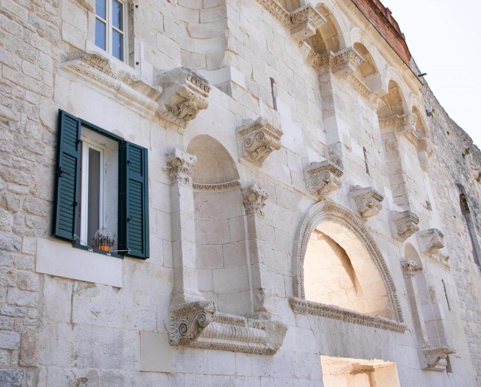 Where to Stay in Split: Hotel & Areas Guide 22 A close-up of an old stone building facade with arched details and ornate decorations—perfect inspiration for those seeking where to stay in Split. An open green shutter window reveals a birdcage, while soft daylight brightens the textured stone.