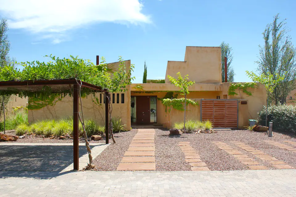 25 Weekend Trips from Madrid Max. 2h Away 15 A modern, flat-roofed house with a stucco exterior, often seen on weekend trips from Madrid, is surrounded by gravel landscaping, a stone pathway, green plants, and a wooden pergola covering part of the driveway under a clear blue sky.