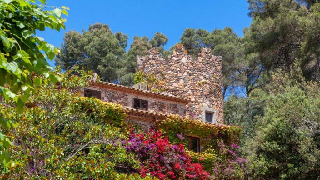 A rustic stone building with a round tower, reminiscent of boutique hotels in Costa Brava, is surrounded by lush greenery and vibrant flowering plants under a clear blue sky, with tall pine trees in the background.