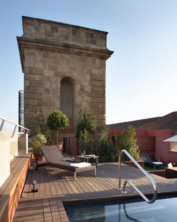 Top Barcelona Hotels with Rooftop Pool 36 A rooftop patio with a small pool, lounge chairs, and lush plants overlooks a historic stone tower under a clear blue sky—one of the charming scenes you’ll find at Barcelona Hotels with Rooftop Pool.