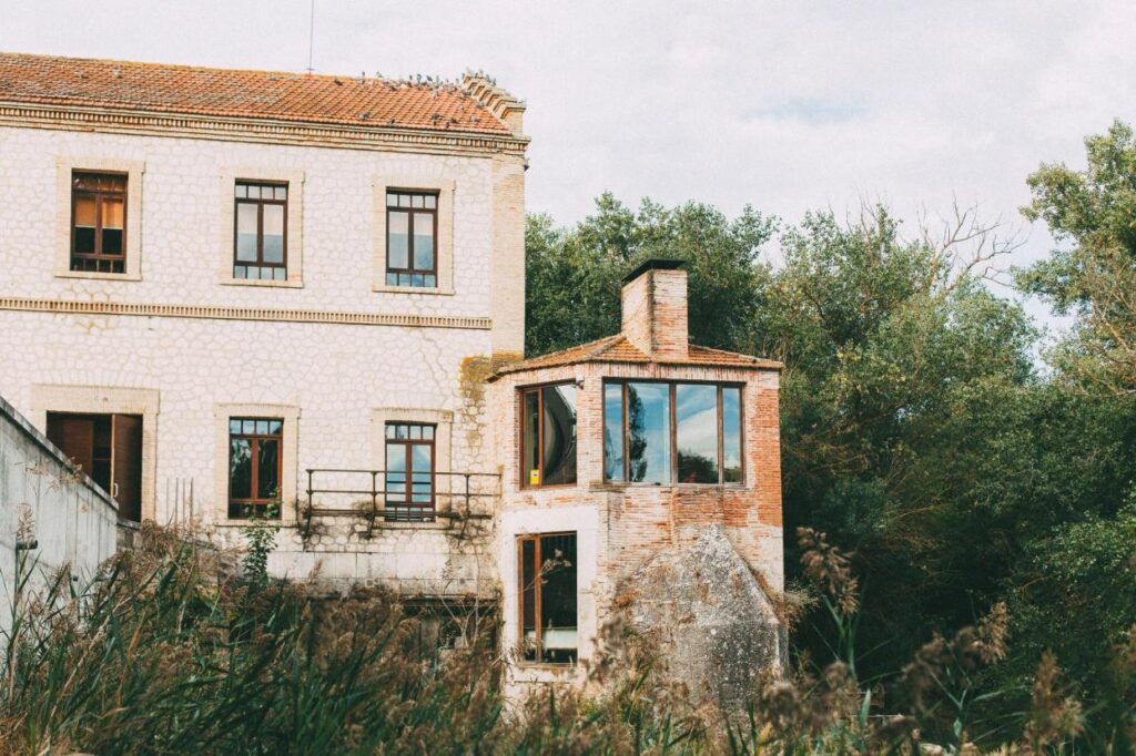 4 Best Hotels in Ribera del Duero, Spain 12 An old stone and brick house with large windows, surrounded by tall grass and dense green trees under a partly cloudy sky—a charming spot often found on weekend trips from Madrid.