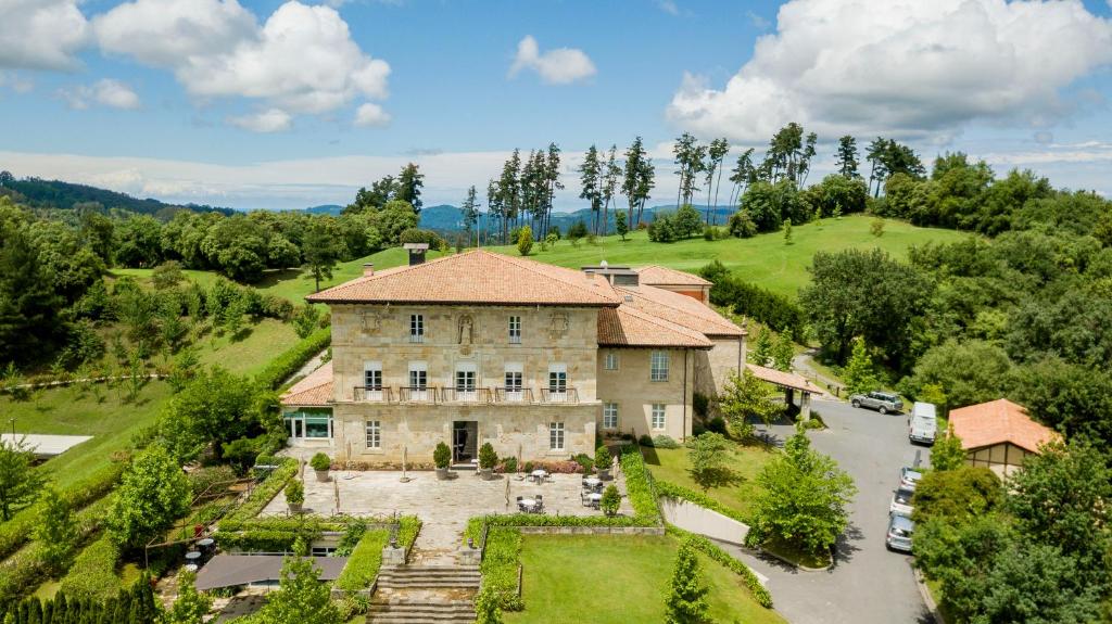 8 Unique Boutique Hotels in Bilbao Spain 18 A large stone country house with a red tile roof, surrounded by green lawns and gardens, evokes the charm of Boutique Hotels in Bilbao. Several cars are parked nearby, with hills and forests visible under a partly cloudy sky.