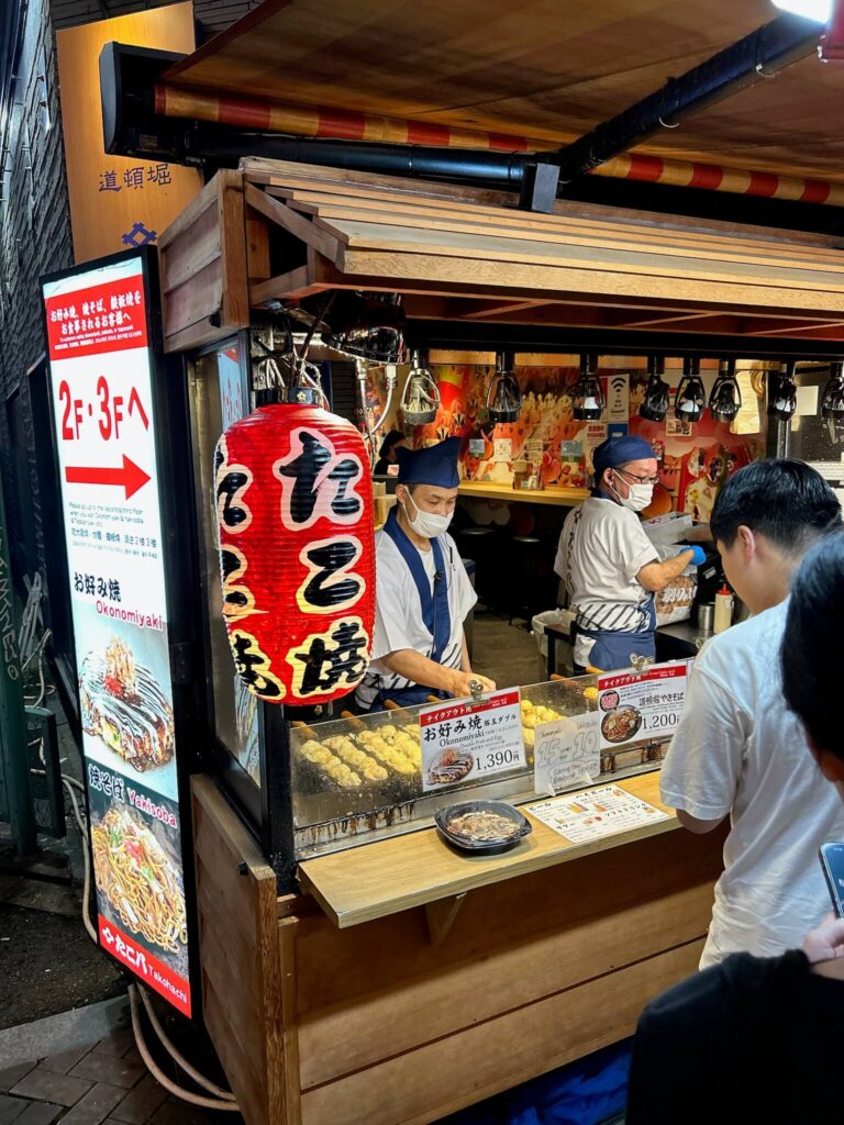 8 BEST Ryokans in Osaka 37 A street food stall in Japan sells takoyaki and okonomiyaki. A chef in a mask and apron prepares food behind the counter, while customers wait in line. Bright signs and a red lantern decorate the stand, popular with visitors from ryokans in Osaka.