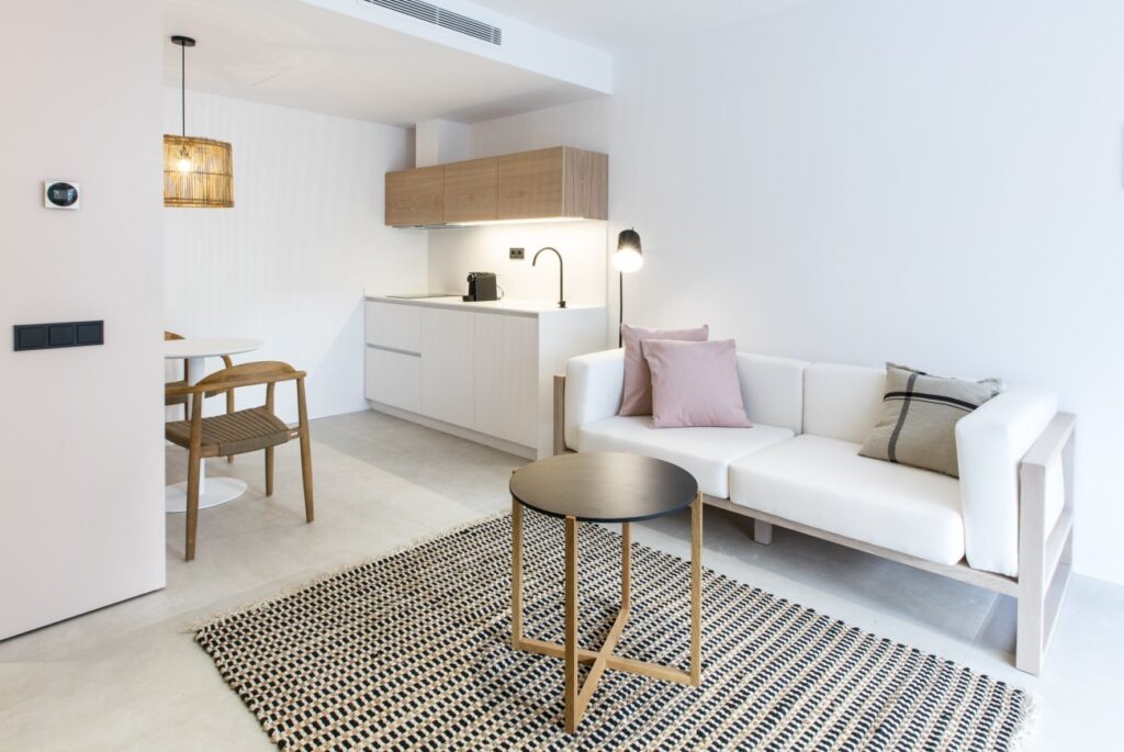 A modern, minimalist living space inspired by boutique hotels in Formentera, featuring a small kitchen, round dining table with wooden chairs, white sofa with pastel cushions, black circular coffee table, and a patterned rug on a light floor.