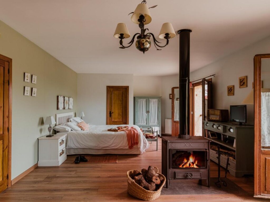 25 Weekend Trips from Madrid Max. 2h Away 77 Cozy bedroom with a double bed, nightstands, and a wood-burning stove—perfect for weekend trips from Madrid. Enjoy warm, rustic decor with wooden flooring, a basket of firewood, TV console, wardrobe, and doors leading outside.