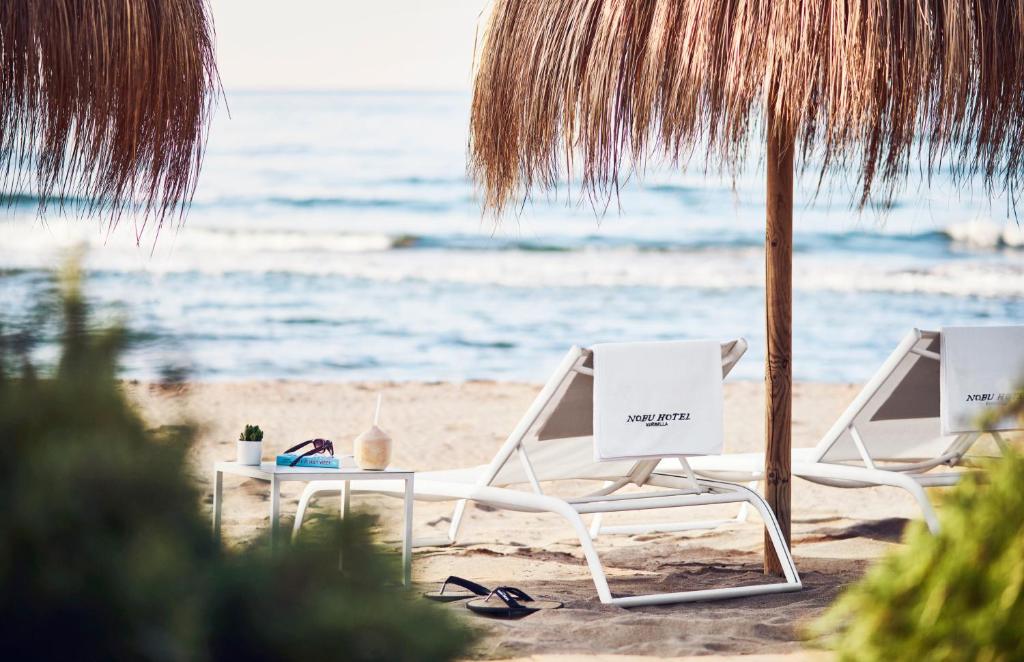 Adults-Only Hotels in Costa del Sol 13 Two white lounge chairs under a thatched umbrella face the sea on a sandy beach at one of the adult only hotels in Costa del Sol. A small table holds drinks and sunglasses, with waves and lush greenery setting a tranquil scene.