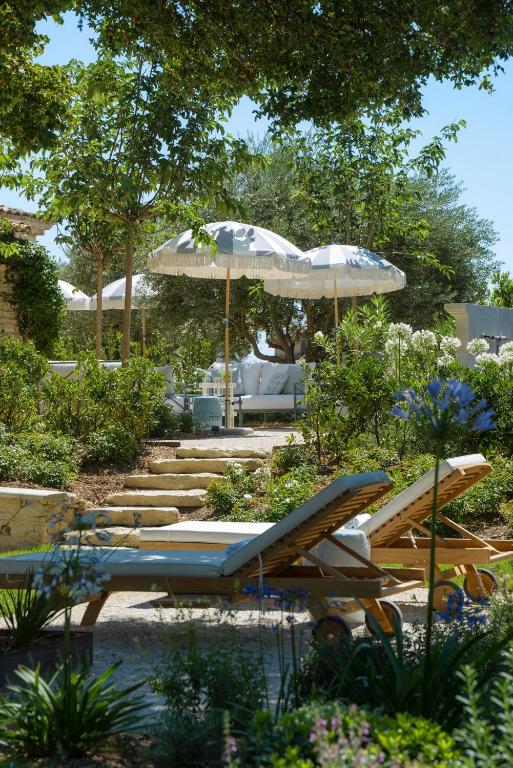 11 Unique Places to Stay in Provence 27 Two wooden lounge chairs with white cushions sit in a lush garden—one of the unique places to stay in Provence. Stone steps lead up to white umbrellas and more seating, all shaded by trees and surrounded by greenery and flowering plants.