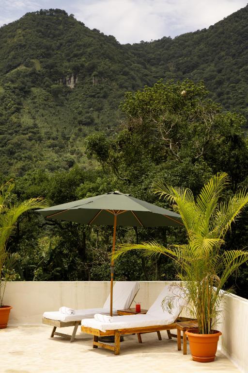 8 UNIQUE Places Where to Stay in Lake Atitlán 21 Two lounge chairs with white cushions sit under a large green umbrella on a terrace, surrounded by potted palm plants, offering one of the most serene views for those seeking where to stay in Lake Atitlan. Lush green mountains and trees complete the backdrop.