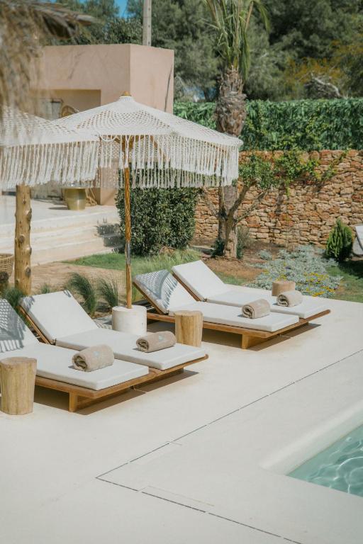 Two poolside lounge chairs with rolled towels sit under a fringed white umbrella beside a swimming pool at one of the boutique hotels in Formentera, surrounded by greenery, a stone wall, and a modern building in the background.