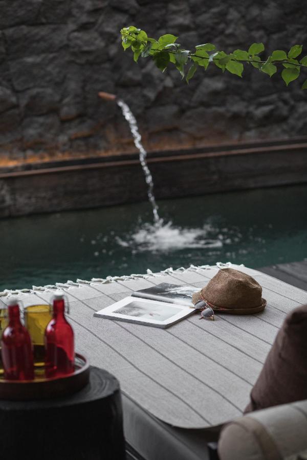 14 UNIQUE Boutique Hotels in Canggu, Bali 2 A cozy outdoor lounge at one of the boutique hotels in Canggu features a gray cushion, a hat, a magazine, and red and yellow bottles on a tray beside a stone wall and a small pool with a water spout, partially shaded by green leaves.