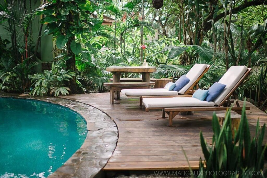 Unique Hotels in Costa Rica: Top Stays 7 A tropical poolside scene at one of the unique hotels in Costa Rica, with two lounge chairs and blue cushions on a wooden deck, beside a round table with benches, all surrounded by lush green plants and trees.