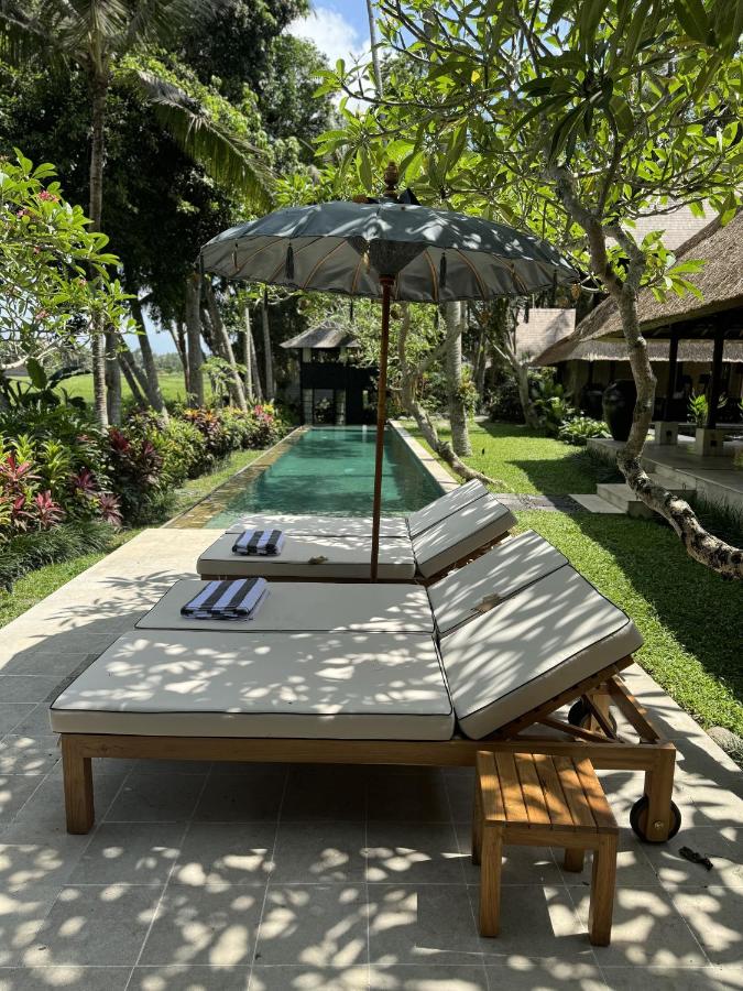 Two cushioned lounge chairs with striped towels sit by a narrow pool, shaded by a large umbrella. Lush tropical plants surround the area, creating the perfect oasis at one of the best family villas in Bali. A thatched-roof building rests to the right.
