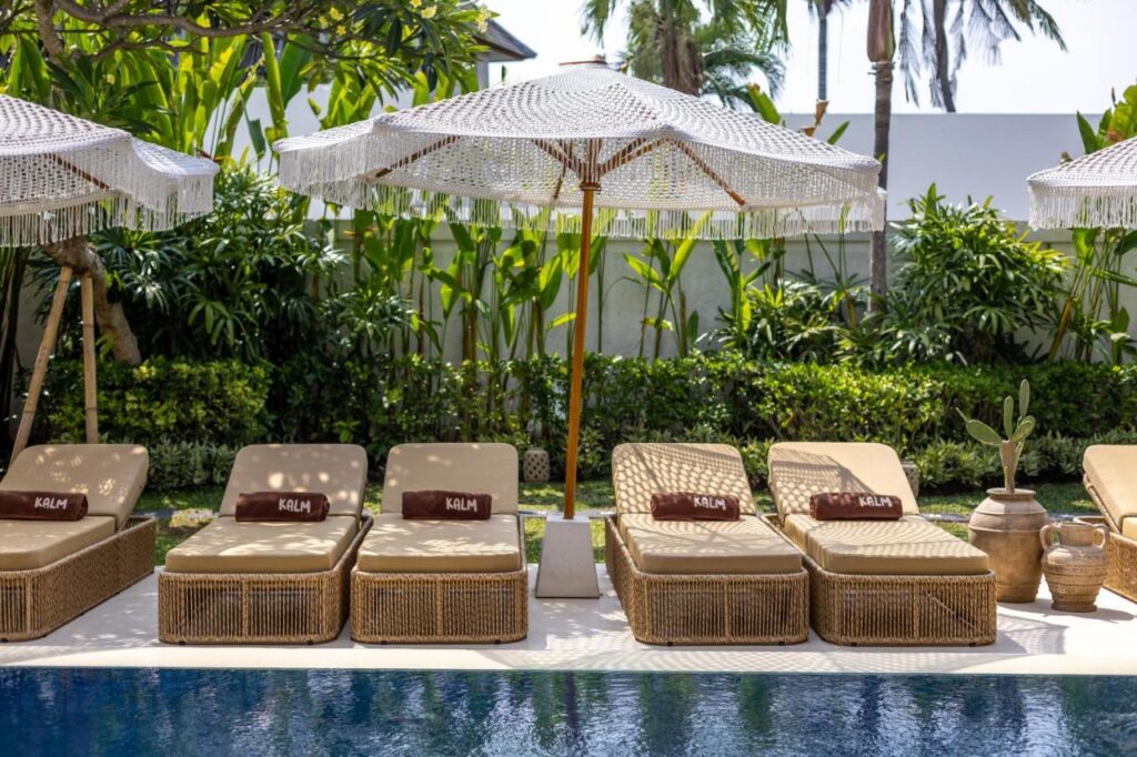 14 UNIQUE Boutique Hotels in Canggu, Bali 35 Four wicker lounge chairs with tan cushions and brown towels reading KALAH line up by a pool, shaded by a white fringe umbrella. Tropical plants surround this relaxing outdoor setting, capturing the vibe of boutique hotels in Canggu.