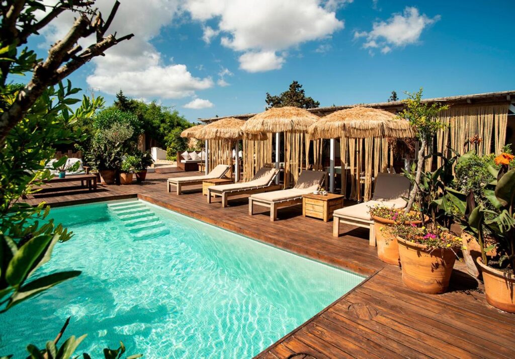 A sunlit outdoor pool with clear water, surrounded by wooden decking, potted plants, and lounge chairs shaded by thatched umbrellas, evokes the serene charm of boutique hotels in Formentera under a partly cloudy sky.