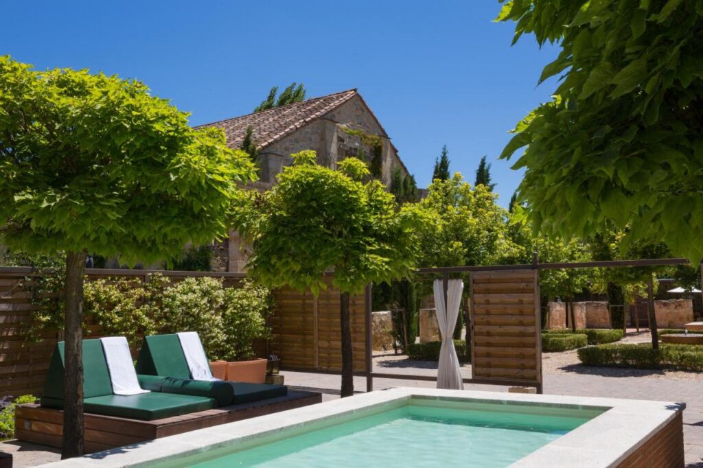 25 Weekend Trips from Madrid Max. 2h Away 24 A small outdoor pool surrounded by wooden lounge chairs with green cushions and towels, shaded by leafy trees—an ideal spot for relaxing during weekend trips from Madrid, with a rustic stone building and blue sky in the background.