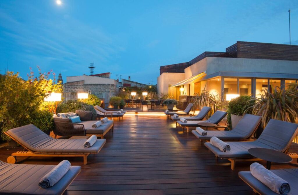 Top Barcelona Hotels with Rooftop Pool 17 A rooftop terrace at dusk in one of the Barcelona Hotels with Rooftop Pool features wooden lounge chairs with rolled towels, small side tables, warm lighting, and lush plants, creating a relaxing and inviting atmosphere.