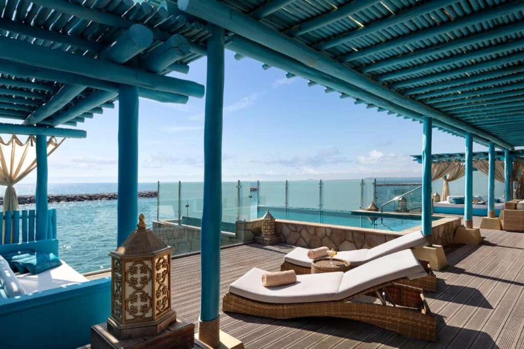 Where to Stay in Doha, Qatar: Transit, City & Beach Hotels 15 A luxurious seaside terrace—perfect for those seeking where to stay in Doha—with turquoise wooden beams, lounge chairs with towels, wicker furniture, glass railings, and a pool overlooking the ocean under a clear blue sky.