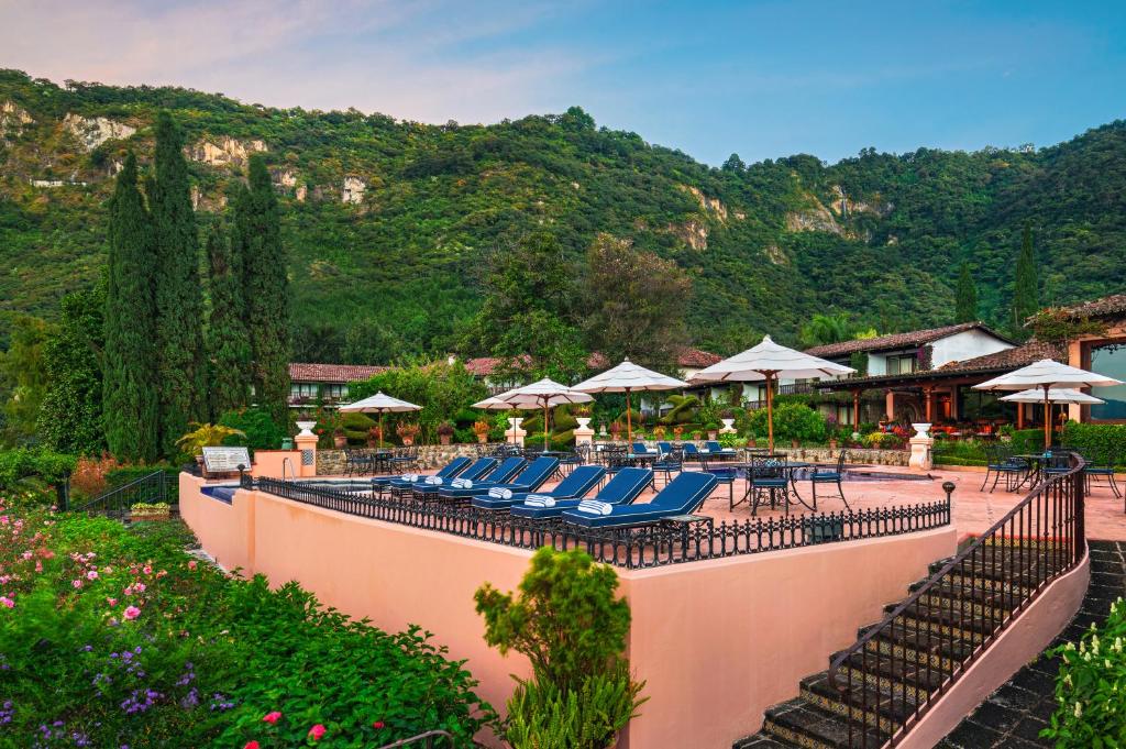 8 UNIQUE Places Where to Stay in Lake Atitlán 31 A terrace with blue lounge chairs and white umbrellas overlooks lush gardens, set against green hills and rustic buildings—an inviting spot for those wondering where to stay in Lake Atitlan under a clear sky.
