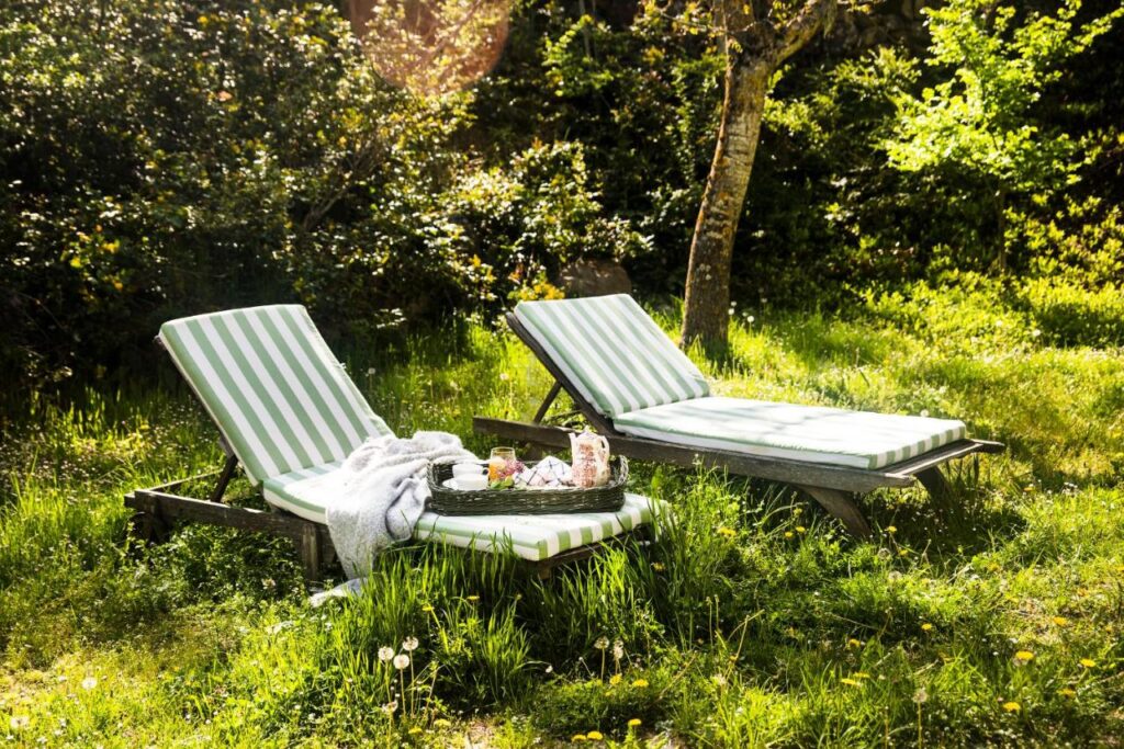 25 Weekend Trips from Madrid Max. 2h Away 66 Two green-and-white striped lounge chairs sit in a sunny, grassy garden next to a small tray with drinks and snacks—perfect for relaxing after weekend trips from Madrid. Sunlight filters through the trees, creating a peaceful and inviting outdoor scene.