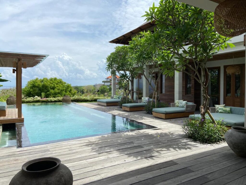 A luxurious outdoor pool surrounded by wooden decking, lounge chairs, and lush greenery beside a modern villa under a partly cloudy sky—one of the best family villas in Bali.