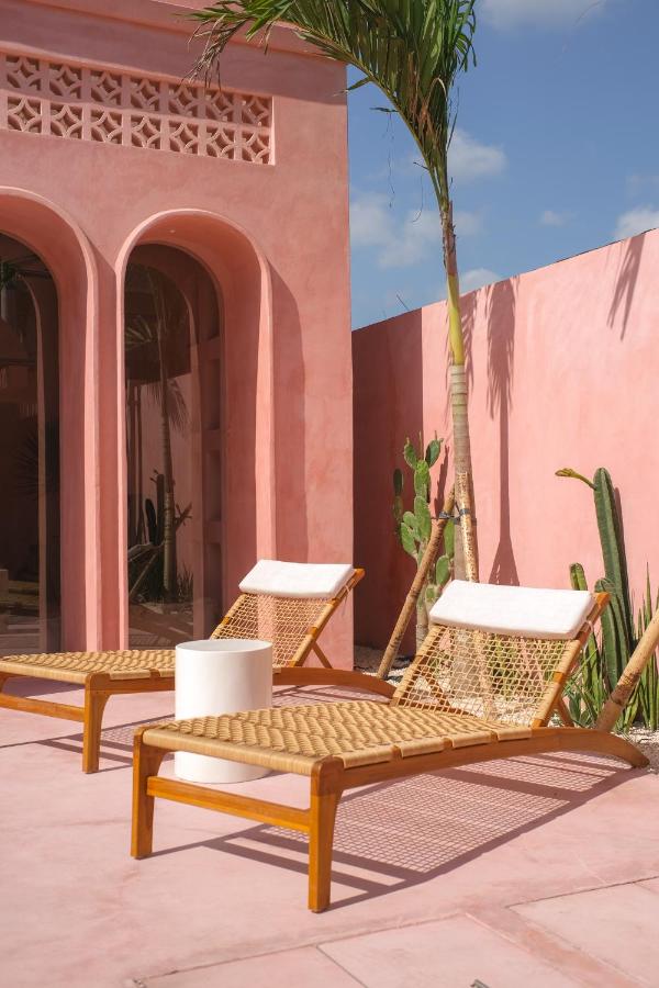 Two wooden lounge chairs with woven seats and white cushions sit on a pink patio beside a pink stucco building, echoing the relaxed style of the best family villas in Bali, with palm trees and cacti set against a clear blue sky.