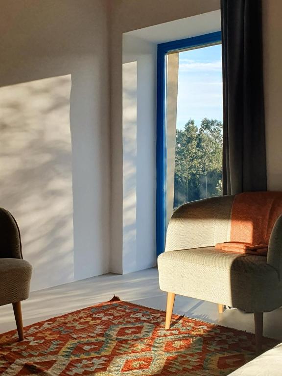 Sunlight streams through a large blue-framed window onto two beige armchairs and an orange patterned rug, creating a serene, modern vibe reminiscent of boutique hotels in Asturias. Trees sway outside as shadows dance on the crisp white walls.