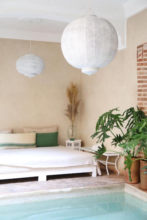 12 Charming Budget Riads in Marrakech 24 A serene indoor space inspired by budget riads in Marrakech, featuring a pool, large white lanterns, a white sofa with green cushions, a plant, and dried grass in a vase against a beige wall with a brick accent.