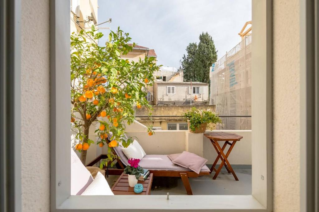 8 TOP Boutique Hotels in Split 12 A view through a window shows a cozy balcony with lounge chair, cushions, small table, potted flowers, and an orange tree full of fruit—just the kind of spot you’d find when searching for where to stay in Split, with nearby buildings under a bright sky.