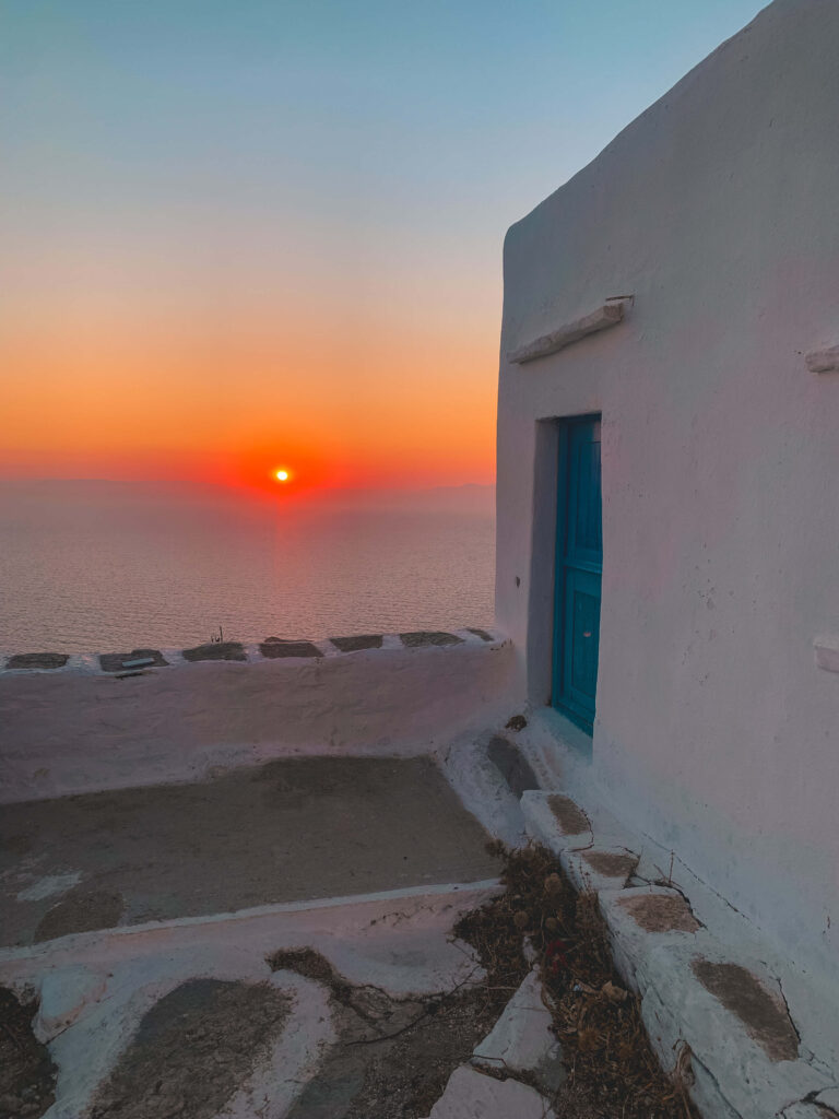A vibrant sunset over the sea, viewed from a whitewashed building with a blue door, characteristic of Greek island architecture. The sky glows orange and pink above the tranquil hotels in Sikinos, casting warm light on the simple stone terrace.