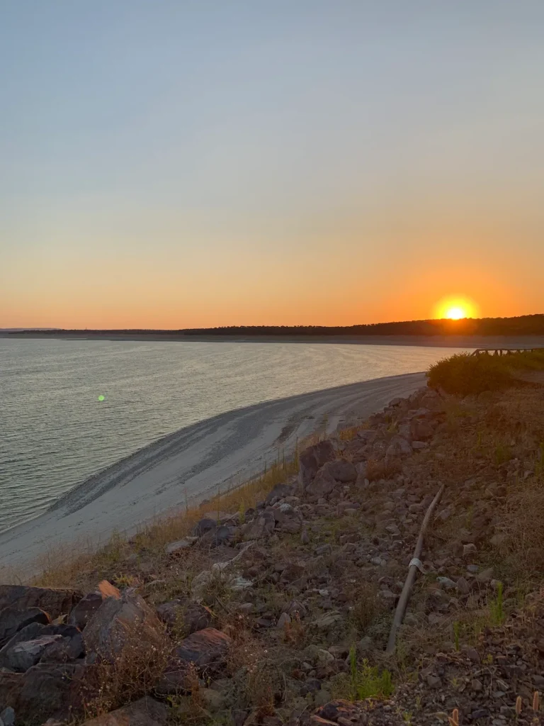 25 Weekend Trips from Madrid Max. 2h Away 17 A serene beach scene at sunset—perfect for weekend trips from Madrid—with the sun low on the horizon, casting a warm orange glow over calm water, sandy shore, and a rocky foreground dotted with greenery.