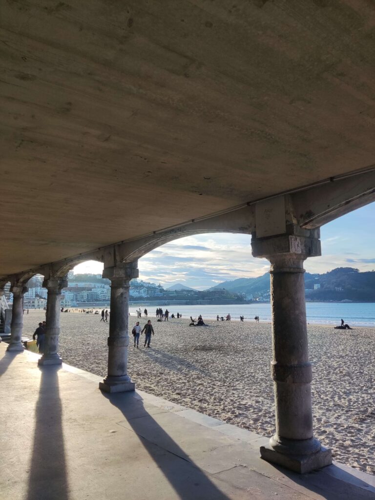 Best Boutique Hotels in San Sebastian 3 People walk and relax on a sandy beach at sunset, viewed from under a concrete arcade with columns. Hills, boutique hotels in San Sebastian, and other buildings are visible in the background under a partly cloudy sky.