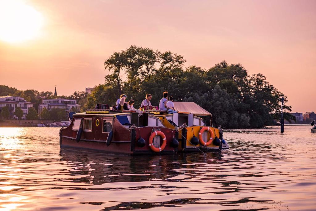 Boutique Hotels in Berlin Kreuzberg 43 A group of people relax on a houseboat on a calm river at sunset, with life rings attached to the boat. In the background, trees and boutique hotels in Berlin Kreuzberg stand under a stunning pink-orange sky.