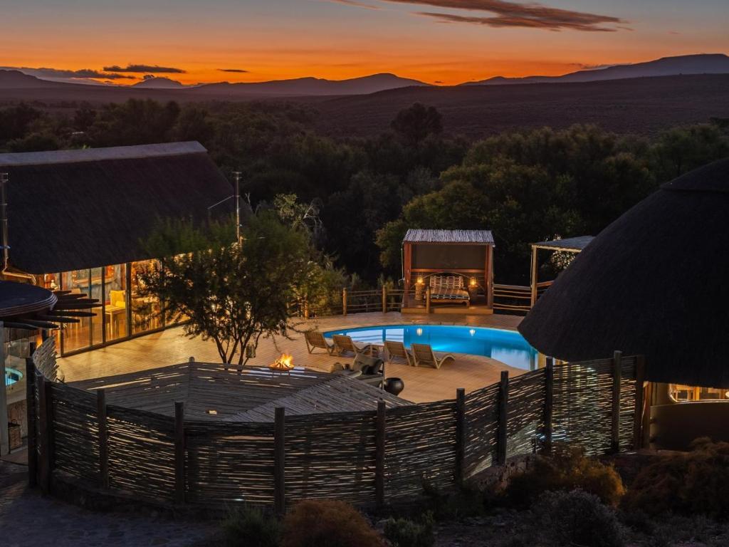 Luxury lodge at sunset with thatched-roof buildings, a glowing outdoor pool, lounge chairs, fire pit, and trees—exemplifying the elegance found at Game Lodges in Western Cape, set against distant hills and an orange sky.