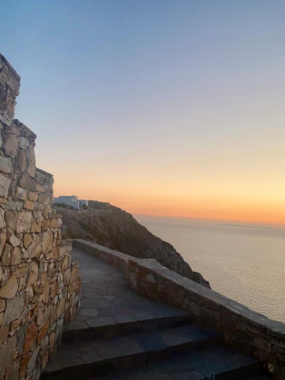 Authentic Hotels in Sikinos, Cyclades 8 Stone pathway along a cliffside overlooking a calm sea at sunset, with pastel colors in the sky and soft light reflecting on the water—just steps away from charming hotels in Sikinos.