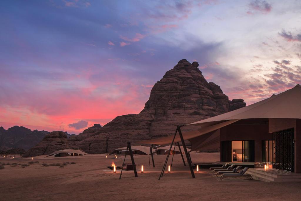 9 Best Hotels in Saudi Arabia: Unique Stays Across the Country 15 Luxury desert tents with lounge chairs are set against dramatic rocky mountains at sunset, offering an inviting atmosphere reminiscent of the Best Hotels in Saudi Arabia, with soft lighting illuminating the vivid pink and purple sky.