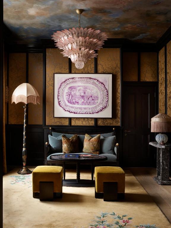 15 Best Boutique Hotels in London, UK 42 Elegant living room reminiscent of boutique hotels in London, featuring a pink feather chandelier, vintage floor lamps with fringed shades, a dark wood sofa with gold pillows, a marble fireplace, and striking framed artwork above the sofa.