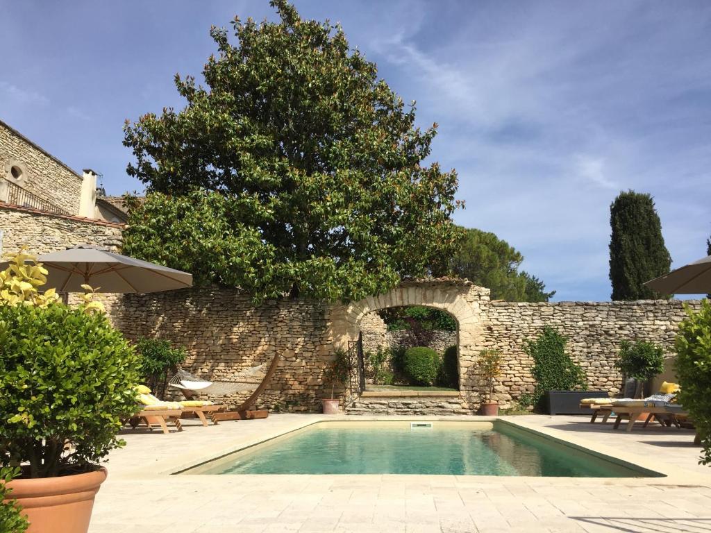 11 Unique Places to Stay in Provence 43 A rectangular swimming pool is surrounded by stone walls, green plants, sun loungers, umbrellas, and a large tree. An arched stone doorway leads to a garden area—one of the unique places to stay in Provence under a blue sky.