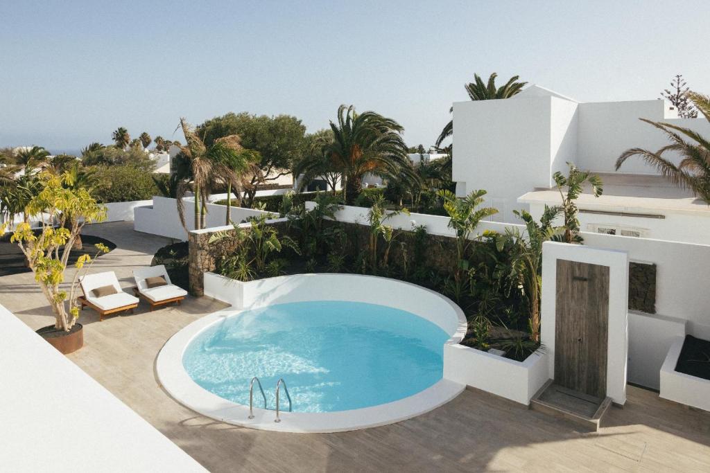 Unique Adults Only Hotels in Lanzarote 19 A modern outdoor space at one of the top Adults Only Hotels in Lanzarote, featuring a circular swimming pool, white buildings, lounge chairs, tropical plants, and palm trees under a clear sky.