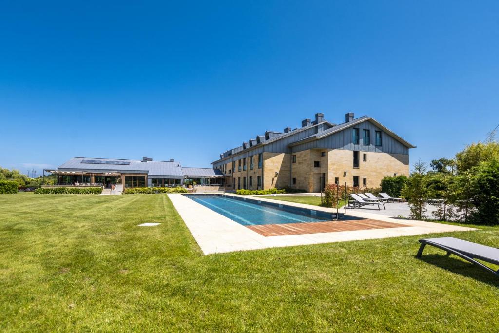 Large modern house with light stone walls and gray roof, next to a rectangular outdoor swimming pool and sun loungers—reminiscent of boutique hotels in Asturias—surrounded by a well-kept green lawn under a clear blue sky.