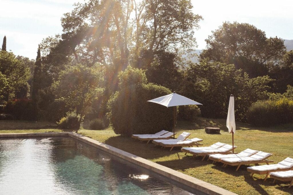 11 Unique Places to Stay in Provence 7 Sunlight streams over a tranquil outdoor pool surrounded by lounge chairs with white cushions and two umbrellas. Lush green trees and bushes fill the background, creating a peaceful garden setting—one of the unique places to stay in Provence.