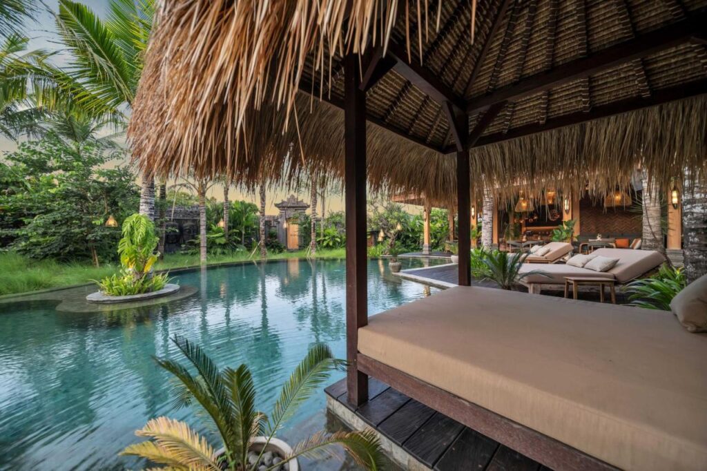 14 UNIQUE Boutique Hotels in Canggu, Bali 9 A tropical resort pool with thatched-roof cabanas and sun loungers, surrounded by lush greenery and palm trees—like the serene escapes found at boutique hotels in Canggu. The pool’s island of plants adds to the relaxing, luxurious atmosphere.