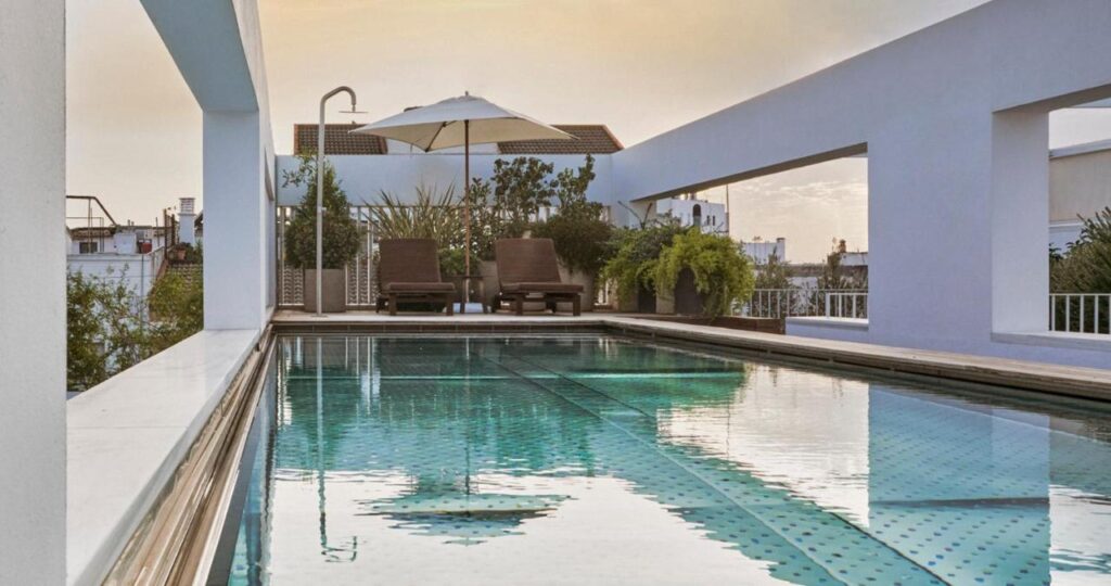 10 UNIQUE Hotels with Pool in Seville 14 A modern rooftop pool with clear water, surrounded by white walls. Two brown lounge chairs and a white umbrella sit at the far end, with green plants nearby—a perfect spot among hotels with pool in Seville, overlooking city buildings and sky.