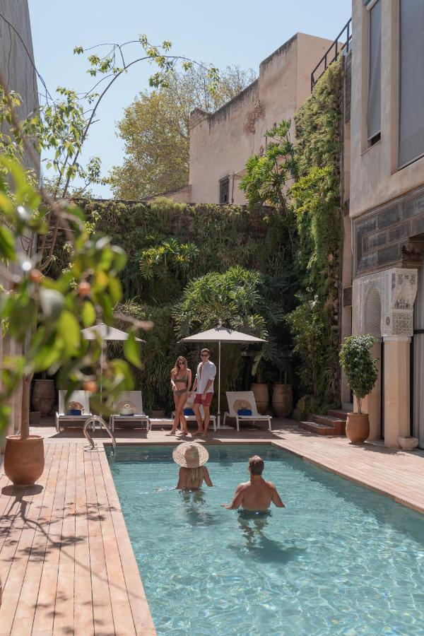 7 UNIQUE Riads in Fez You Won't Forget 17 Four people relax by a rectangular outdoor pool surrounded by greenery, reminiscent of riads in Fez. Two stand under an umbrella, while two others enjoy the tranquil water between tall building walls on a sunny day.