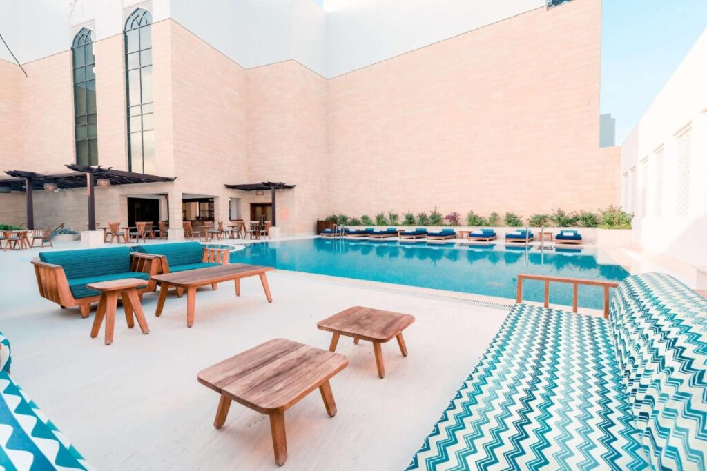 Where to Stay in Doha, Qatar: Transit, City & Beach Hotels 42 Modern outdoor pool area with blue and white patterned lounge chairs, wooden tables, shaded seating, and a row of sunbeds—an inviting spot perfect for those searching where to stay in Doha, surrounded by tall beige walls and large windows.