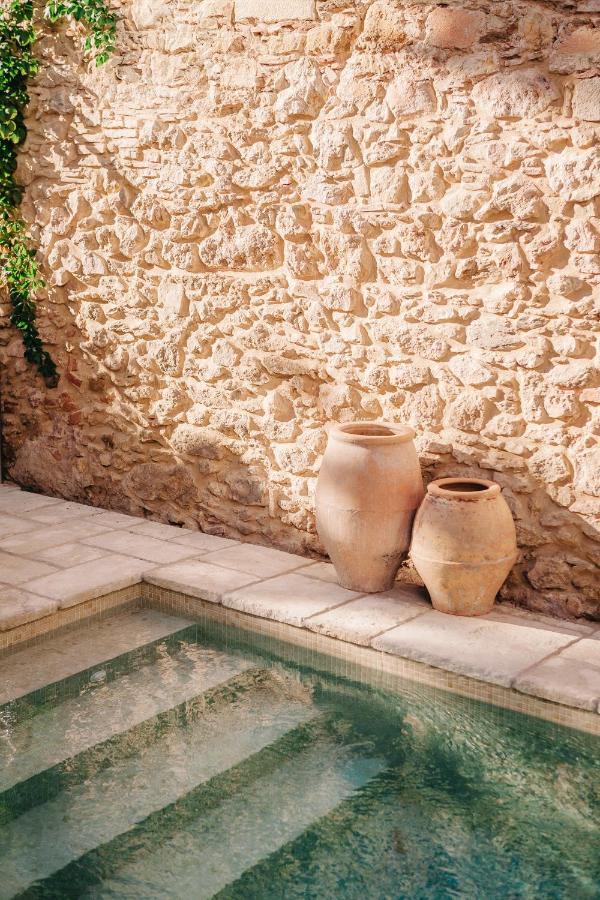 Two large clay pots sit beside a stone wall near a clear, sunlit swimming pool with tiled edges; sunlight casts soft shadows on the rustic scene, evoking the tranquil charm of boutique hotels in Costa Brava.