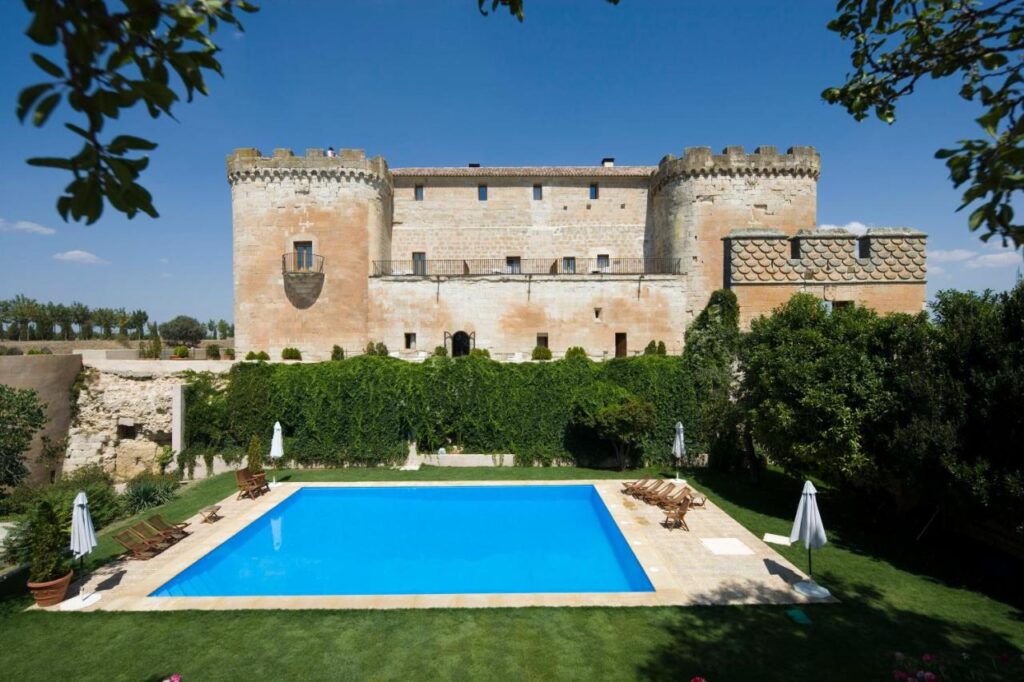 25 Weekend Trips from Madrid Max. 2h Away 26 A historic stone castle with round towers rises behind a rectangular outdoor swimming pool, perfect for weekend trips from Madrid, surrounded by sun loungers, umbrellas, and lush green lawns under a clear blue sky.