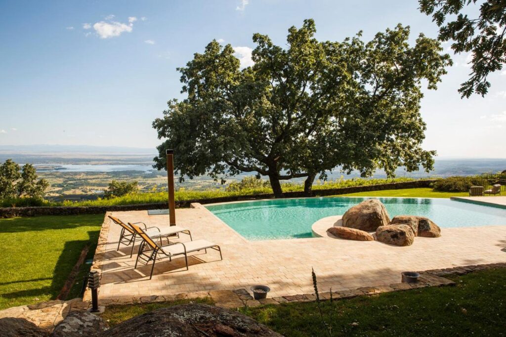 25 Weekend Trips from Madrid Max. 2h Away 41 A serene outdoor pool, perfect for weekend trips from Madrid, features large rocks and lounge chairs on a stone patio shaded by a grand tree. The pool overlooks a lush green landscape and distant hills under a clear blue sky.