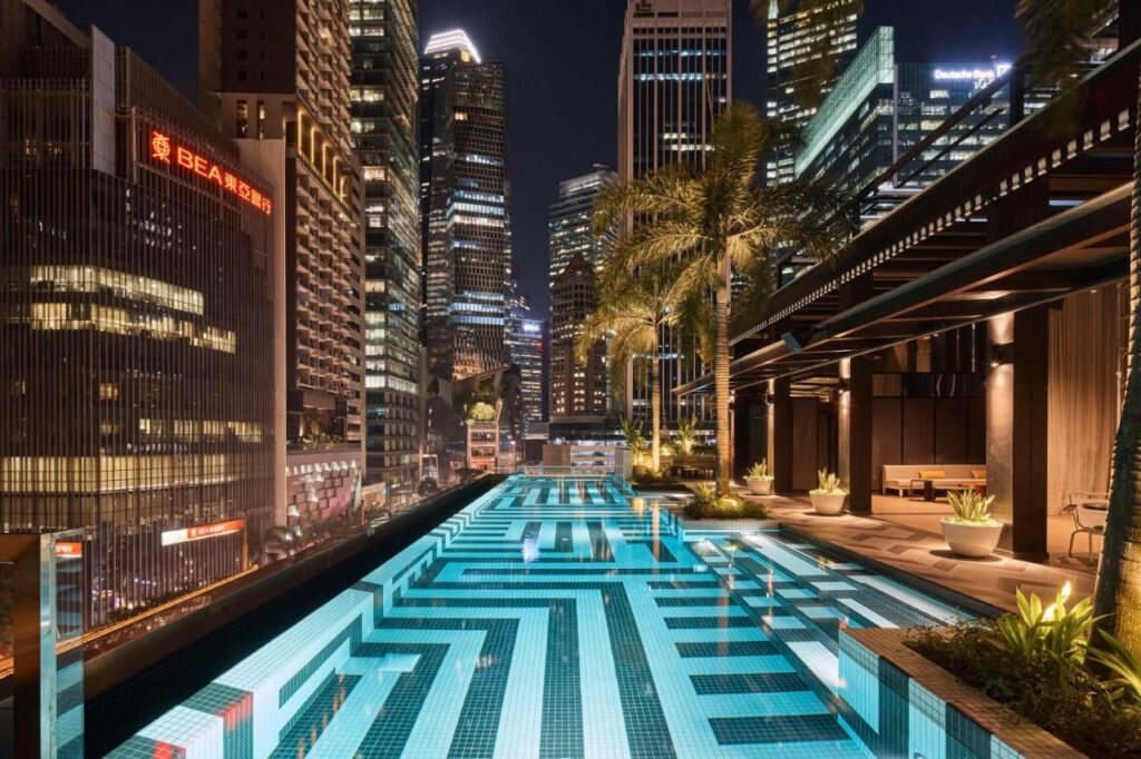 13 Most Unique Hotels in Singapore for an Incredible Holiday 31 A rooftop swimming pool with geometric blue patterns overlooks a cityscape of tall, illuminated skyscrapers at night, with palm trees and modern lounge seating—an experience found at some of the most unique hotels in Singapore.