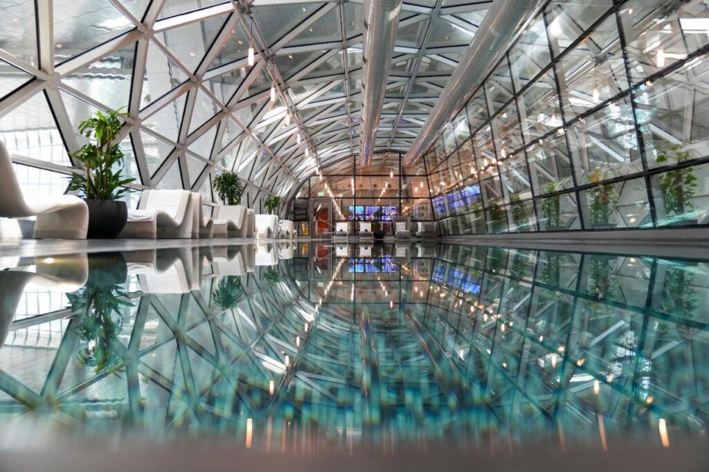 Where to Stay in Doha, Qatar: Transit, City & Beach Hotels 21 A modern indoor space, perfect for those exploring where to stay in Doha, featuring a geometric glass ceiling, lush potted plants, white chairs, and a long reflective pool that beautifully mirrors the elegant architecture and lighting.